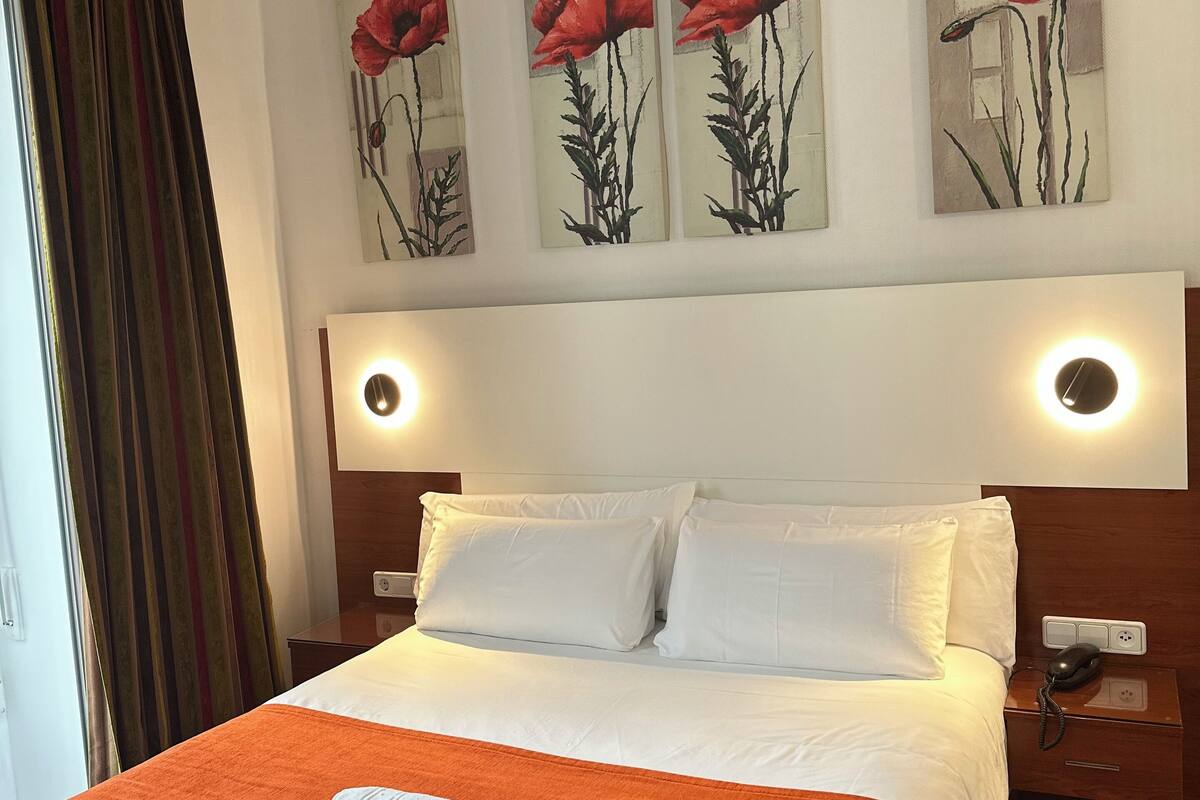 A hotel room with a large bed, white bedding, an orange throw blanket, and three red poppy paintings on the wall.