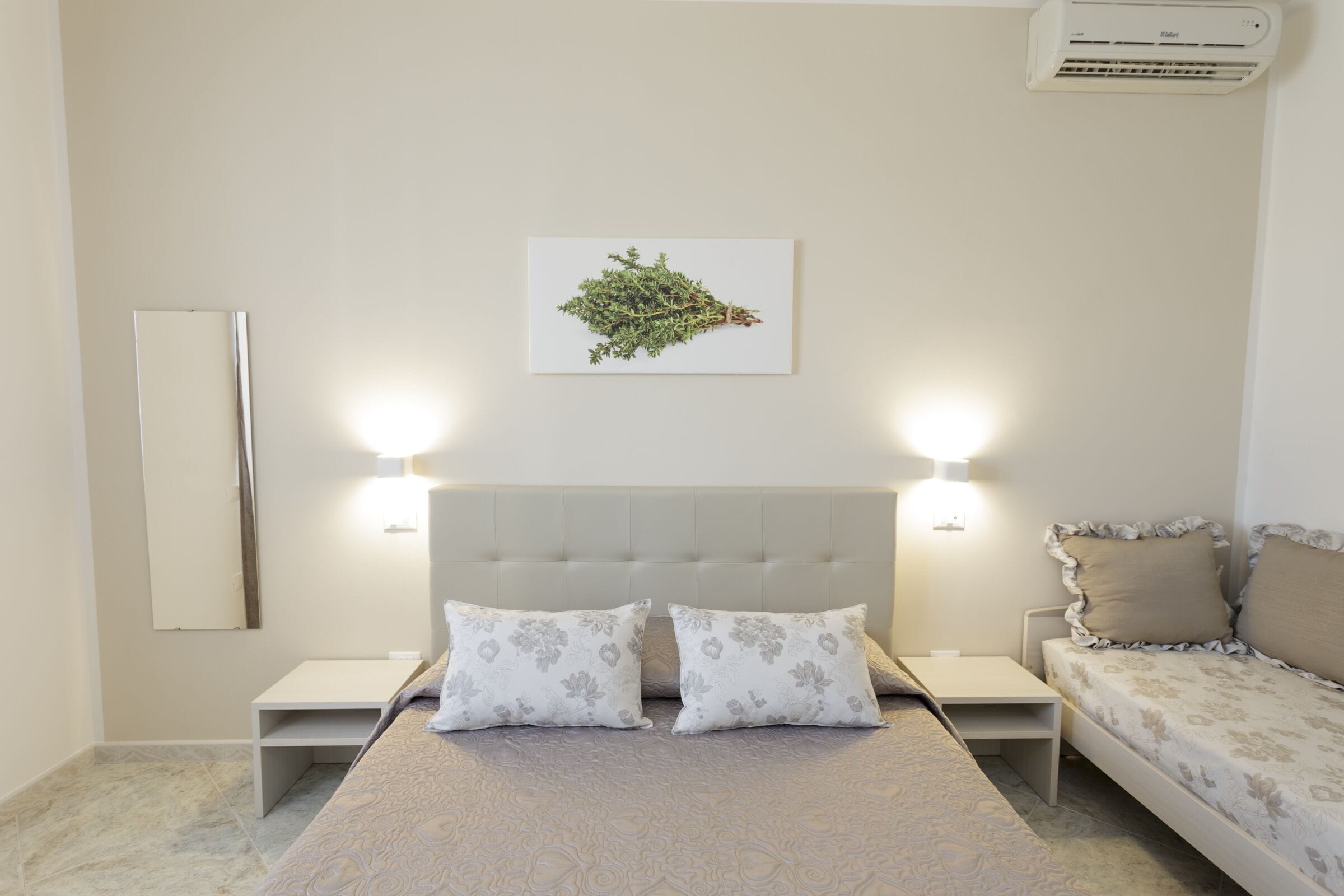 Un dormitorio con cama, dos mesitas de noche, una mesita auxiliar, aire acondicionado mural y un espejo montado en la pared.
