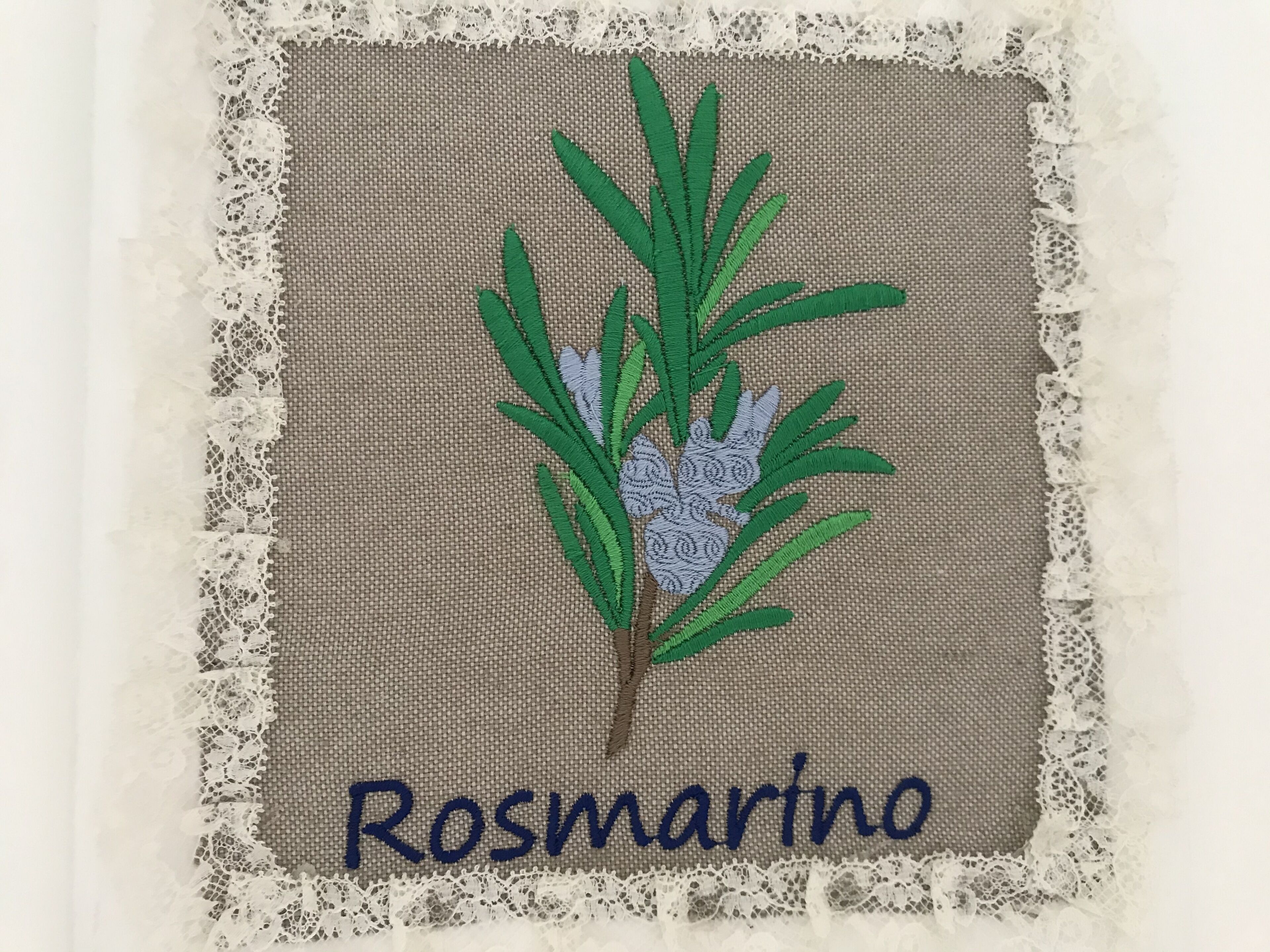 A square embroidered patch with a pine branch and the name "Rosmarino" on a textured fabric background, surrounded by a lace border.