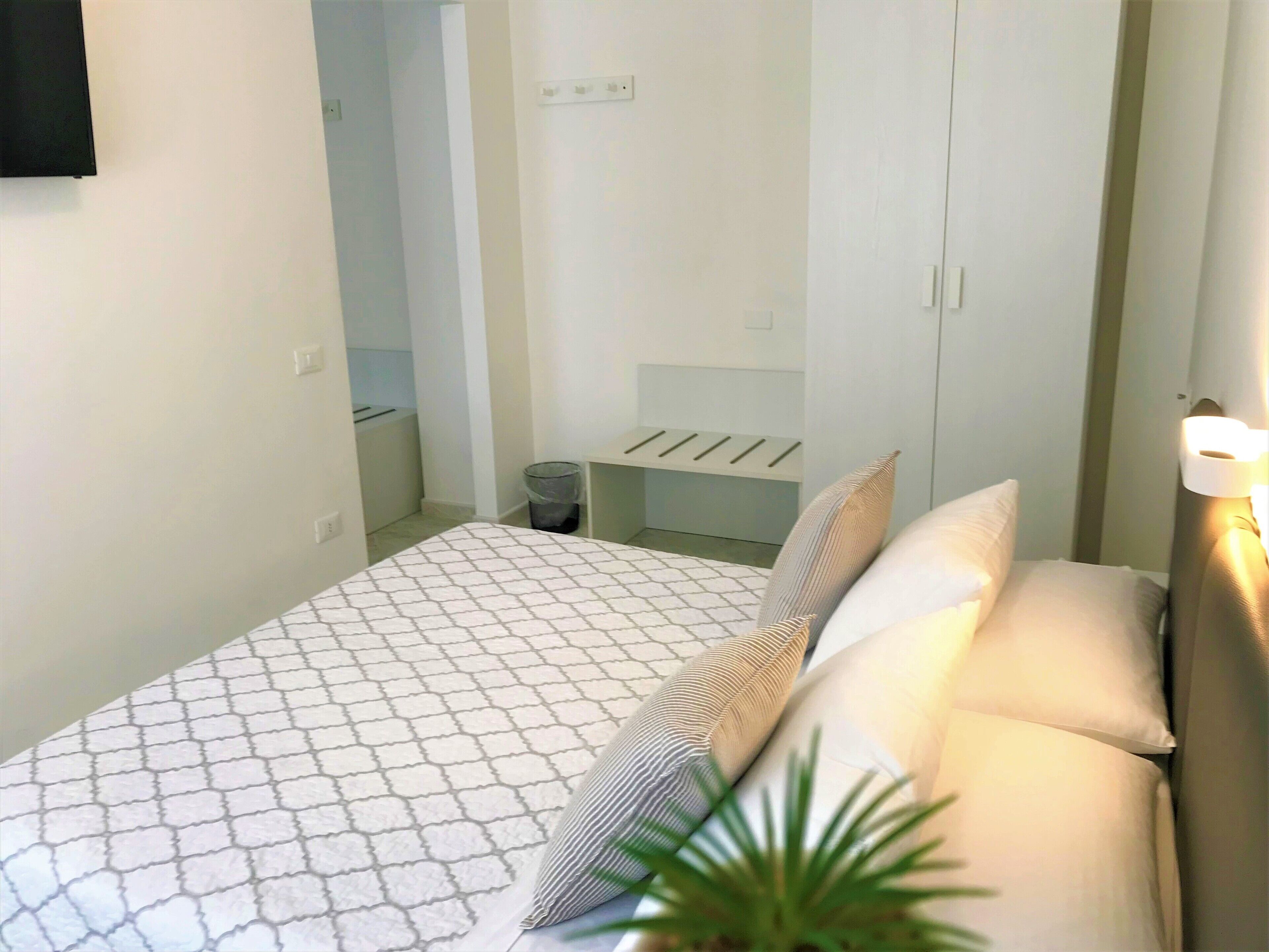 A small, neatly arranged bedroom with a bed, pillows, a bedside table, and a wall-mounted light.