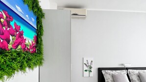 Une chambre avec un lit, une table de chevet, un climatiseur fixé au mur, un tableau encadré et une fresque murale colorée représentant des éléments de verdure.