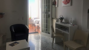 Une pièce dotée d’une porte-fenêtre coulissante donnant sur un espace extérieur aménagé avec une table et des chaises. On y trouve une étagère blanche sur laquelle sont posés un panier et une bouteille.