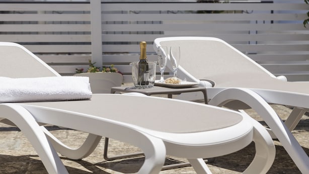 Un espace extérieur ensoleillé, aménagé avec des chaises longues blanches et une table où sont disposés une bouteille, des verres et une assiette de nourriture.