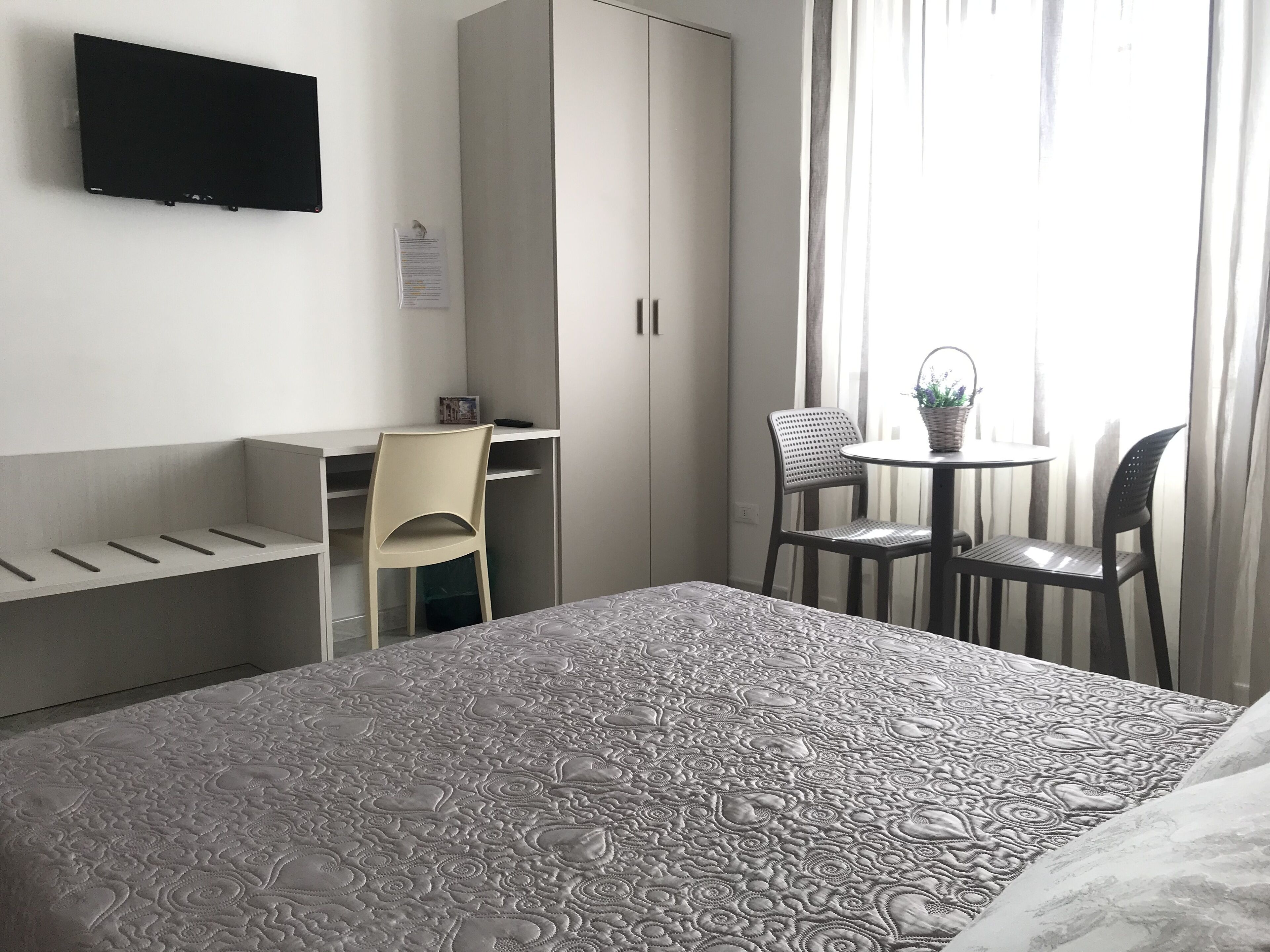 Ett hotellrum med en säng, ett skrivbord, en stol, en tv som är monterad på väggen och ett fönster med gardiner.