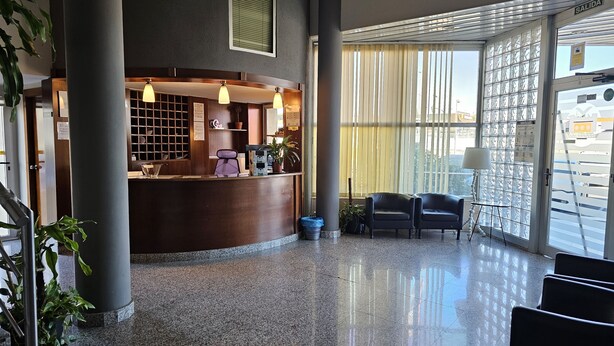A reception area with a wooden desk, two chairs, a potted plant, and a large window with blinds.