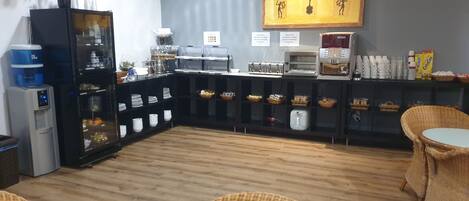 Cafetería con barra de desayuno tipo bufé, que incluye un refrigerador, máquina de café y diferentes opciones de comida.