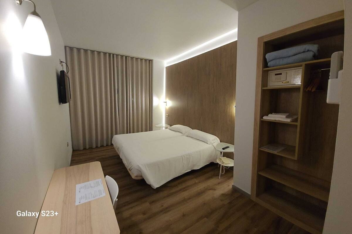 A hotel room with a bed, bedside table, lamp, wardrobe, and a desk with a chair.