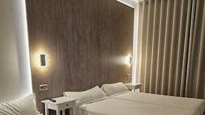 A hotel room with two beds, a small bedside table, a wall-mounted light, and a large window with curtains.