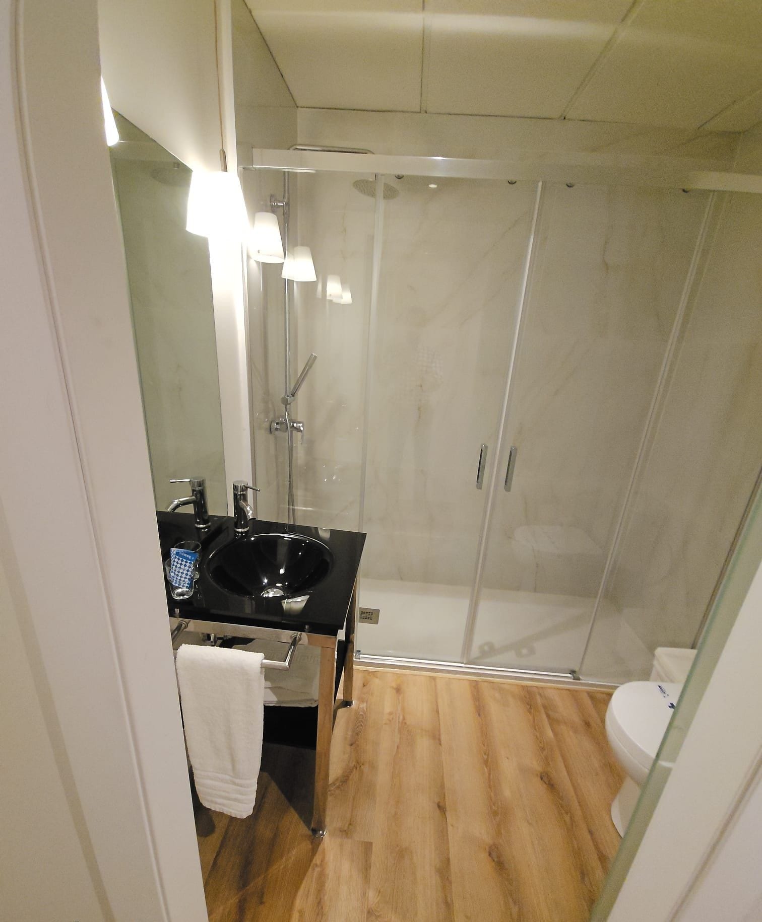 Un baño moderno con ducha de vidrio, un lavabo negro y suelo de madera.