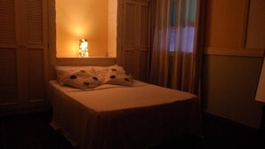 Una habitación tenuemente iluminada con una cama, una ventana con cortinas y un aplique en la pared.