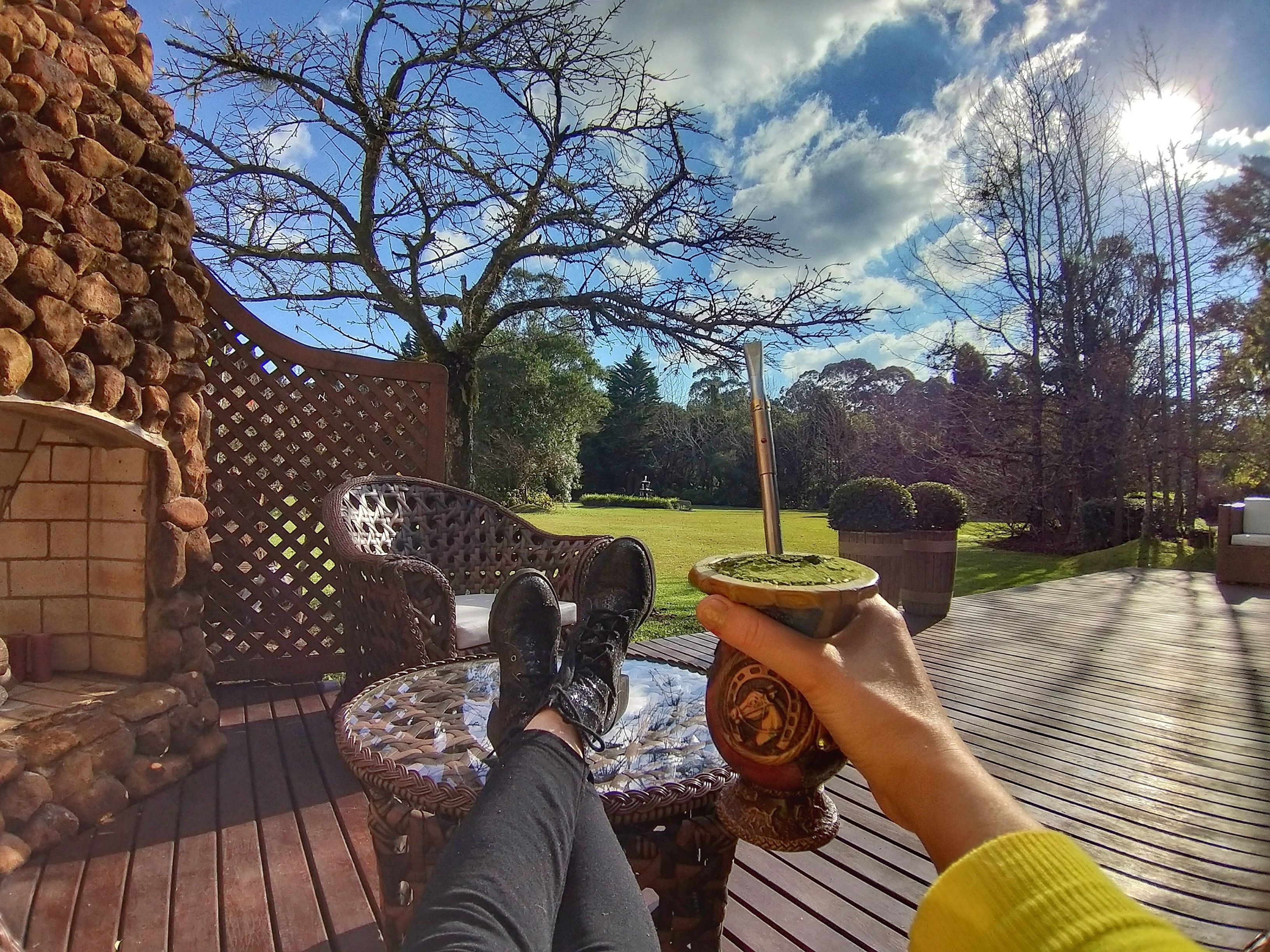 Una persona relajándose en una terraza de madera con un muro de piedra y una silla de mimbre, sosteniendo una taza con un líquido verde.