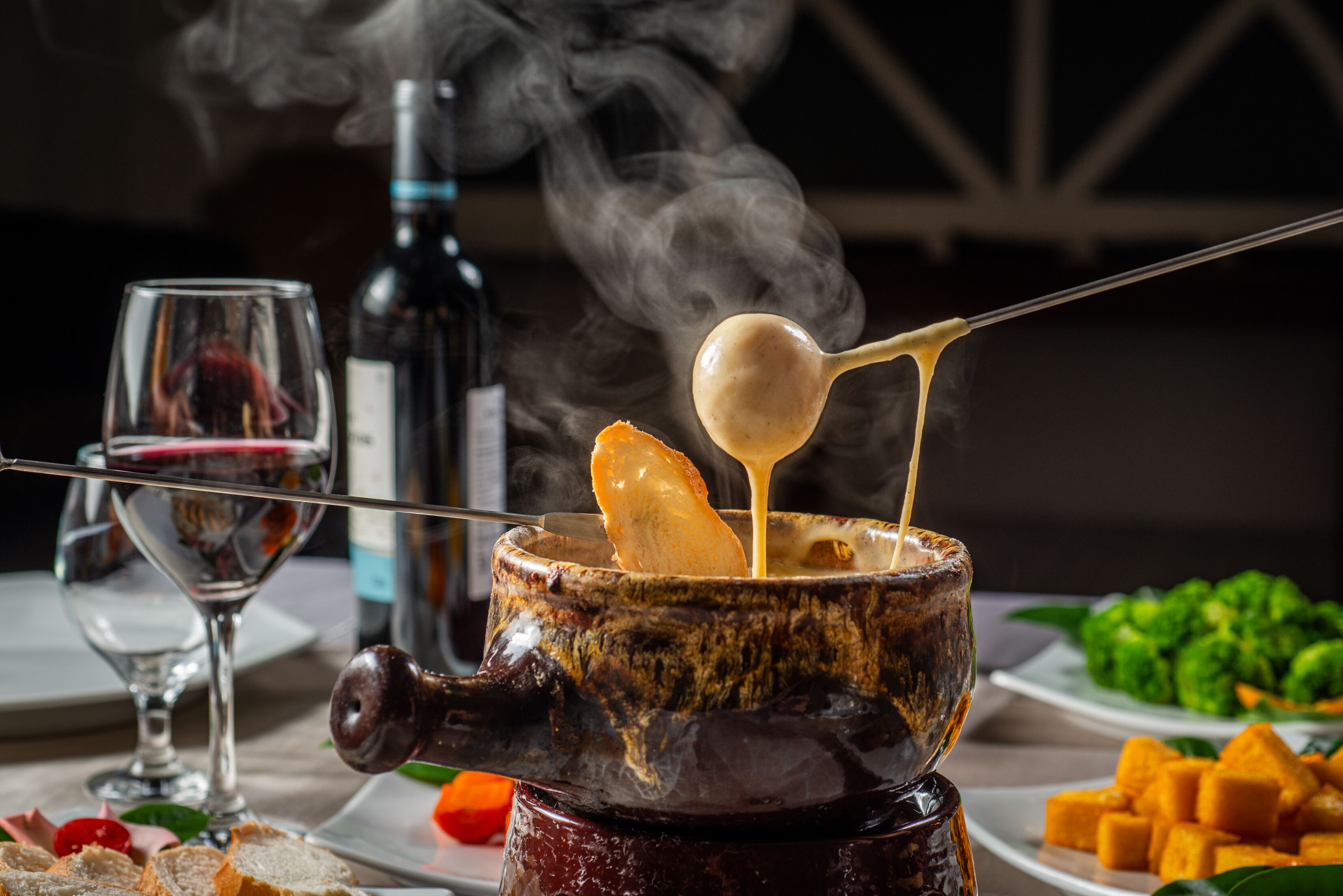 Ein Käsefondue mit einem Brot-Stick zum Dippen, umgeben von Teller mit Essen und einer Weinflasche.