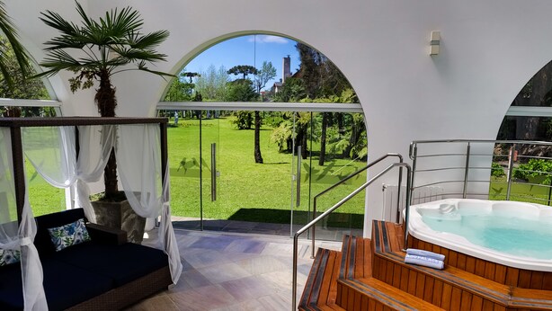A modern outdoor relaxation area with a wooden hot tub, a cushioned seating area with a canopy, and a view of a well-maintained garden.