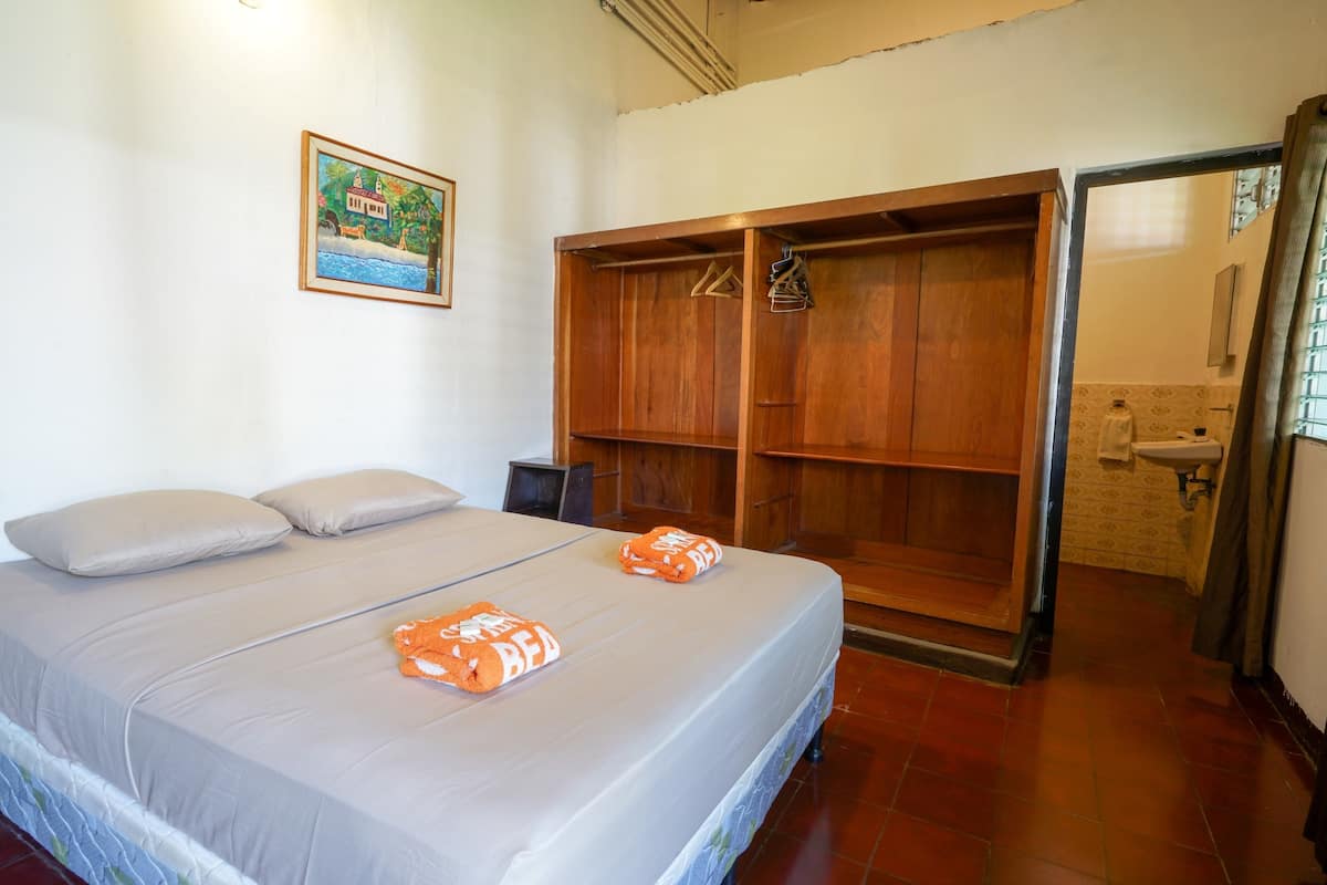 Une chambre à coucher avec un lit, une armoire en bois, une salle de bain visible à travers une porte ouverte et un tableau accroché au mur.
