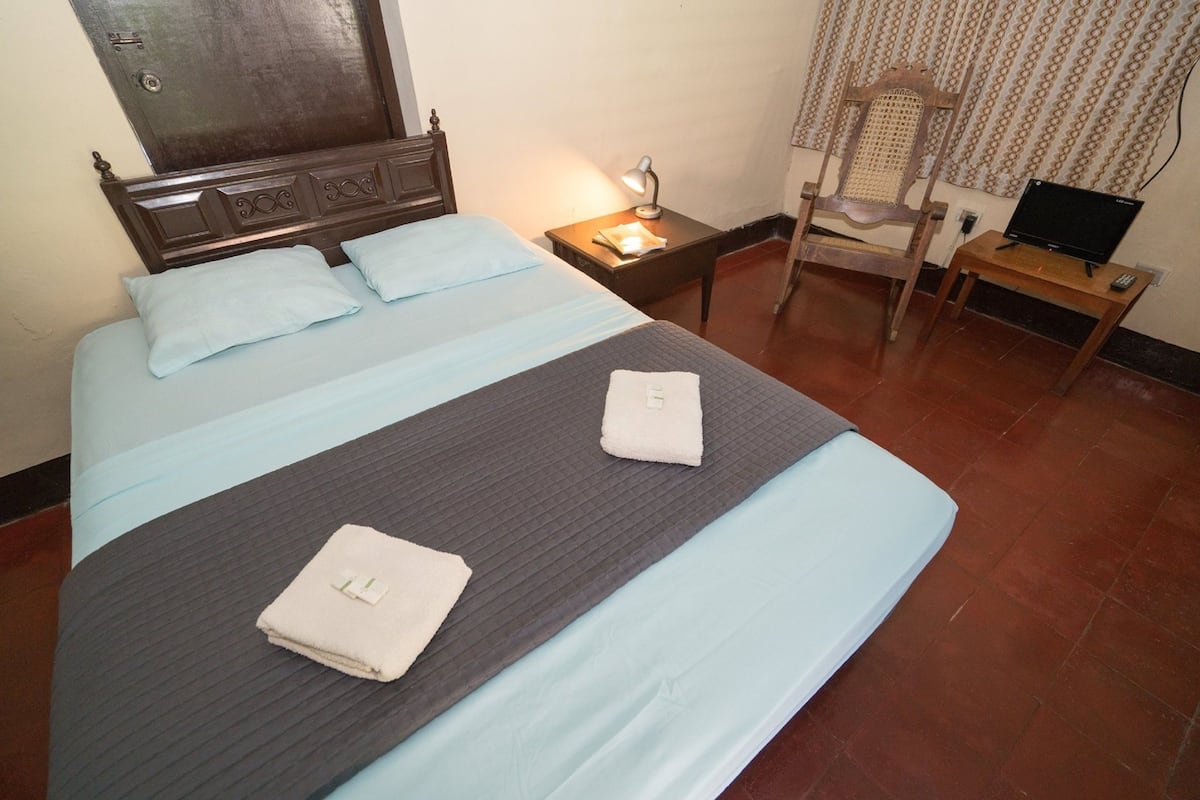 Un lit bien fait, avec des oreillers et une serviette pliée, une lampe de chevet, une chaise et une petite table sur laquelle se trouve un ordinateur portable.