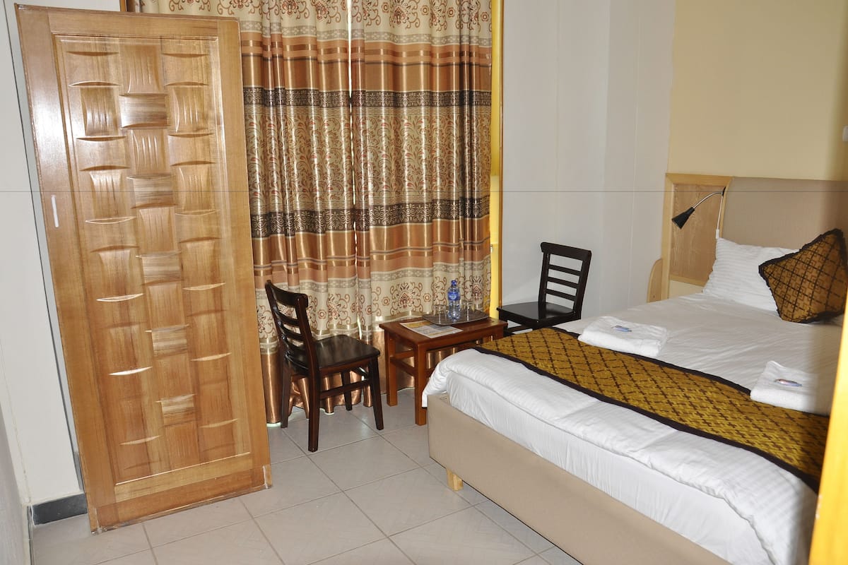 Une chambre d’hôtel avec un lit, un bureau, une chaise, une armoire en bois et un rideau.