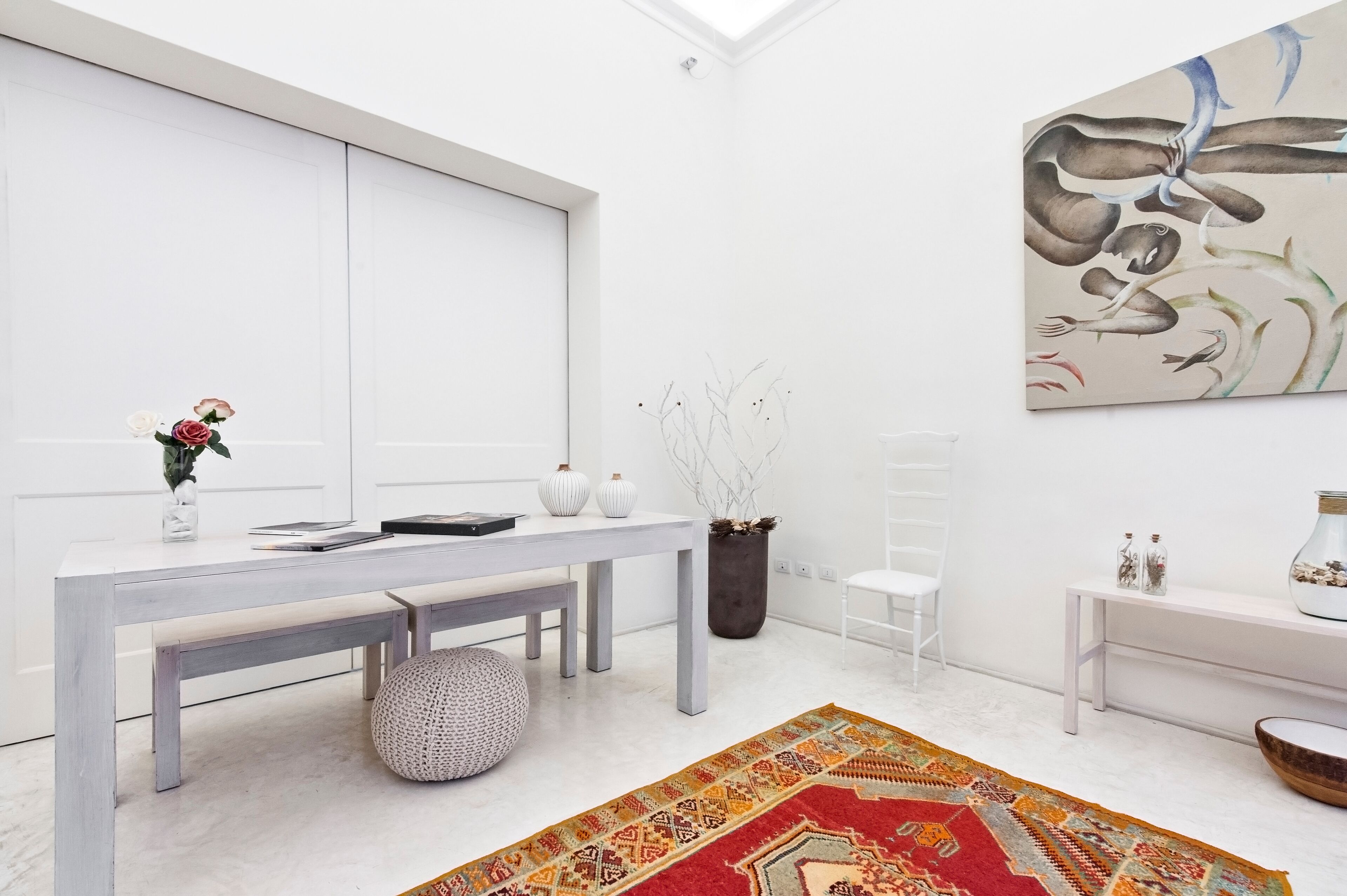 A modern, minimalist room with a white table, bench, and chairs. A colorful rug is on the floor, and a large abstract painting is on the wall.