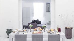 Una mesa de desayuno preparada con diversos alimentos y bebidas, un sofá gris al fondo y una pared blanca con una obra de arte enmarcada.