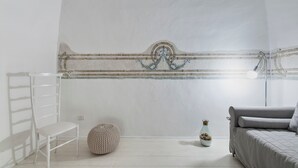Una habitación con una silla blanca, un reposapiés redondo, un jarrón pequeño y un sofá gris.