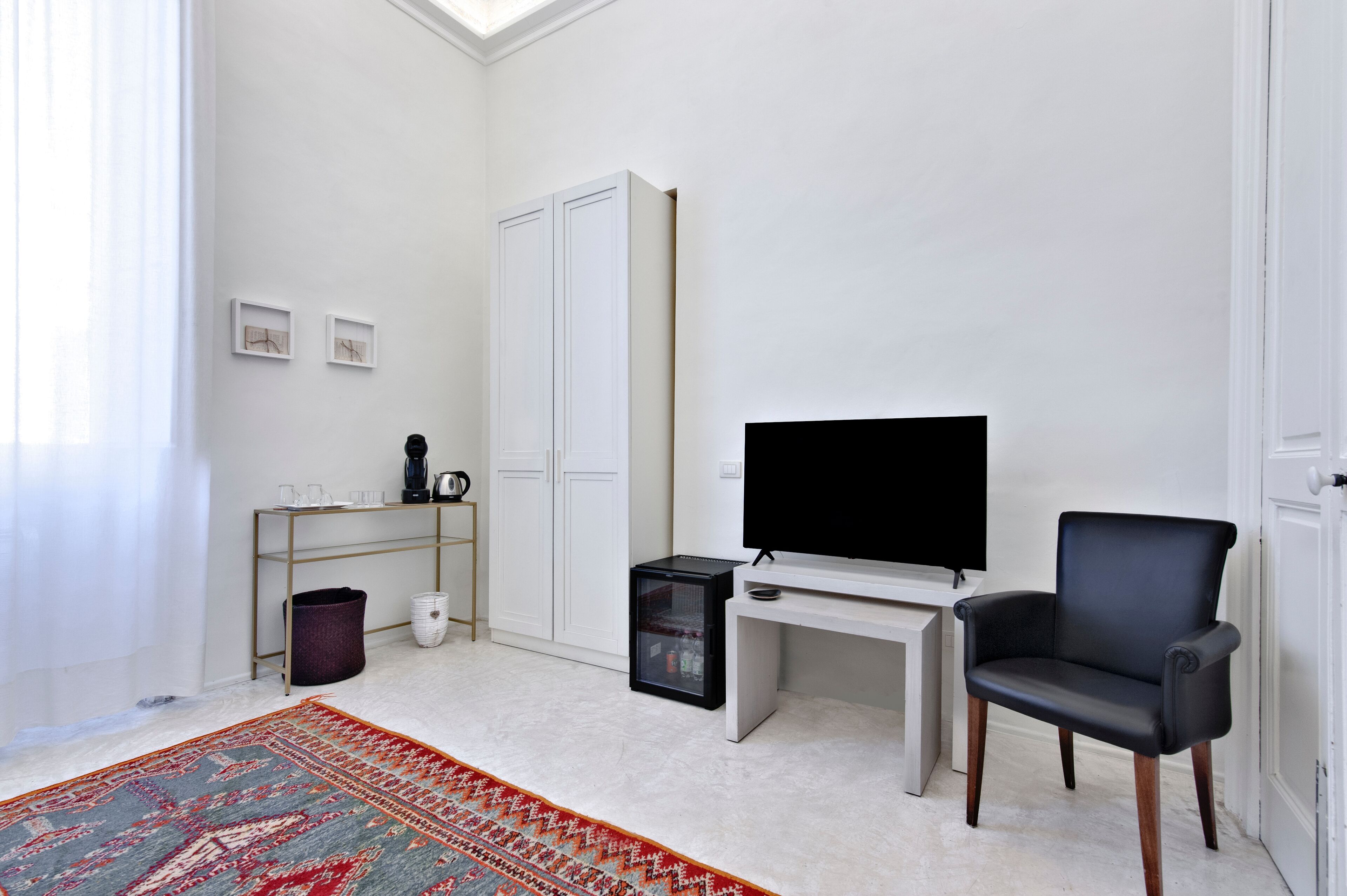 A modern, minimalist room with a large flat-screen TV, a black leather chair, a white desk, and a patterned rug.