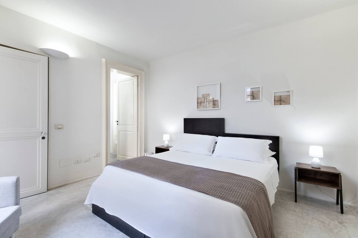 A neatly arranged bedroom with a large bed, bedside tables with lamps, and a door leading to another room.