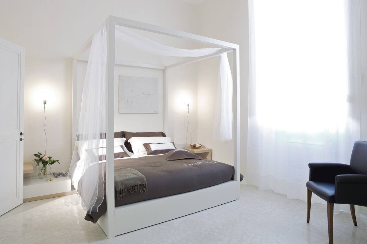 A modern bedroom with a four-poster bed, bedside tables, a nightstand, a chair, and a vase with flowers.