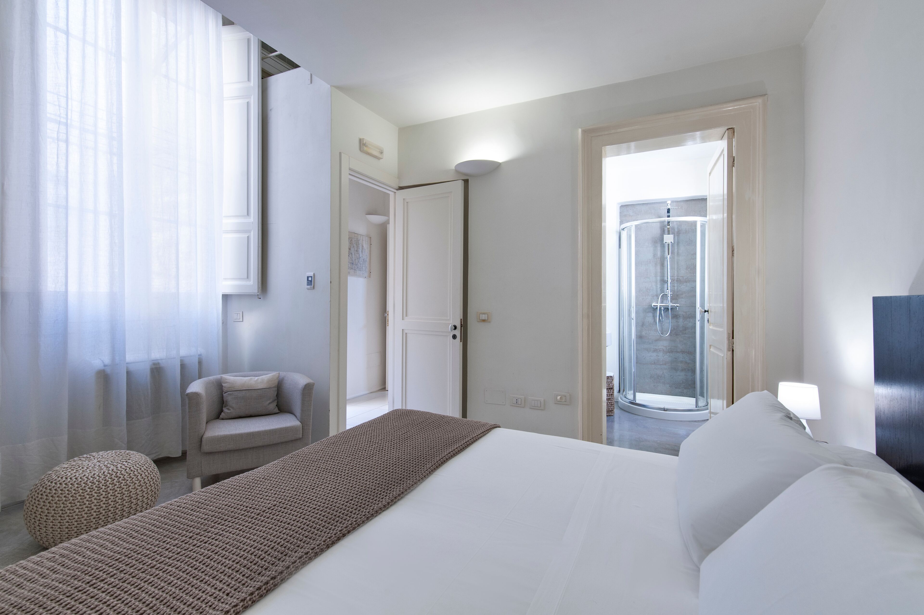A hotel room with a bed, a seating area with a chair, a small ottoman, a door leading to a bathroom, and a shower area.