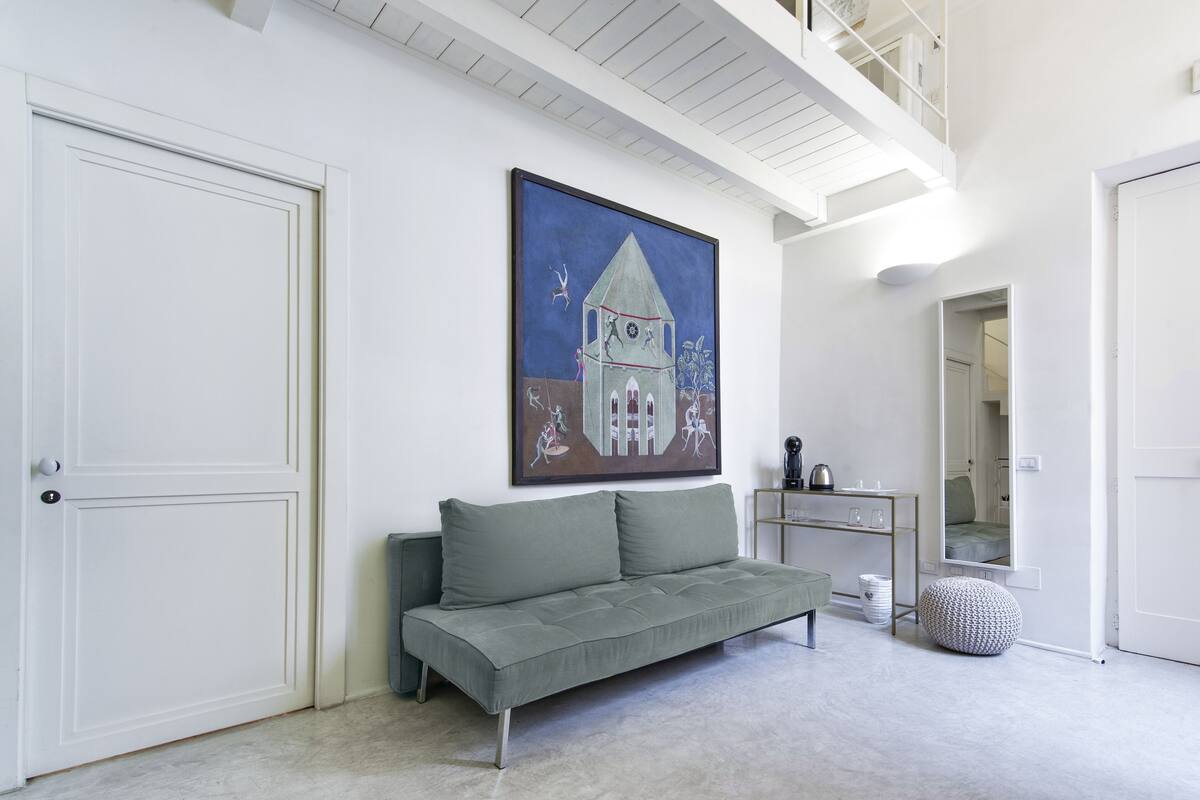 A modern living room with a grey sofa, a painting of a house, a desk with a mirror, and a staircase.