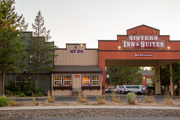The Sisters Inn & Suites entrance with a 'NOW OPEN' sign, a clock, and an American flag.
