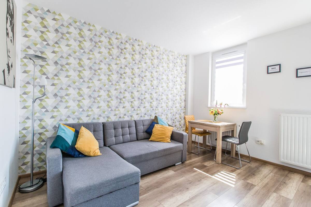 Een moderne woonkamer met een grijze modulaire bank, een houten eettafel en een wand met een geometrisch patroon.