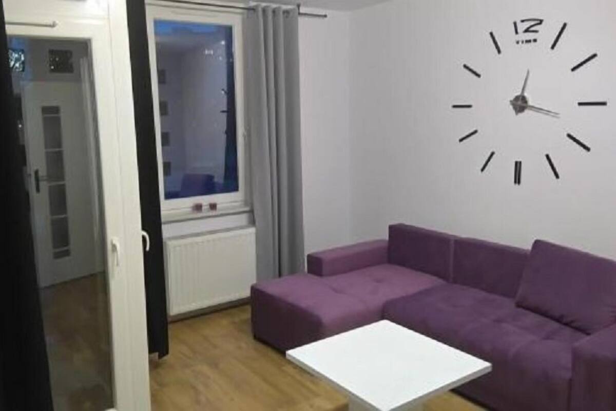 Een woonkamer met een paarse bank, een witte salontafel en een wandklok.