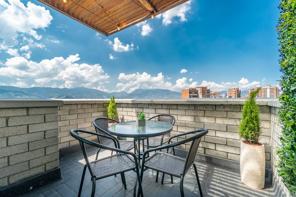 En tagterrasse med et rundt bord og stole, en potteplante og udsigt til fjerne bygninger og bjerge.