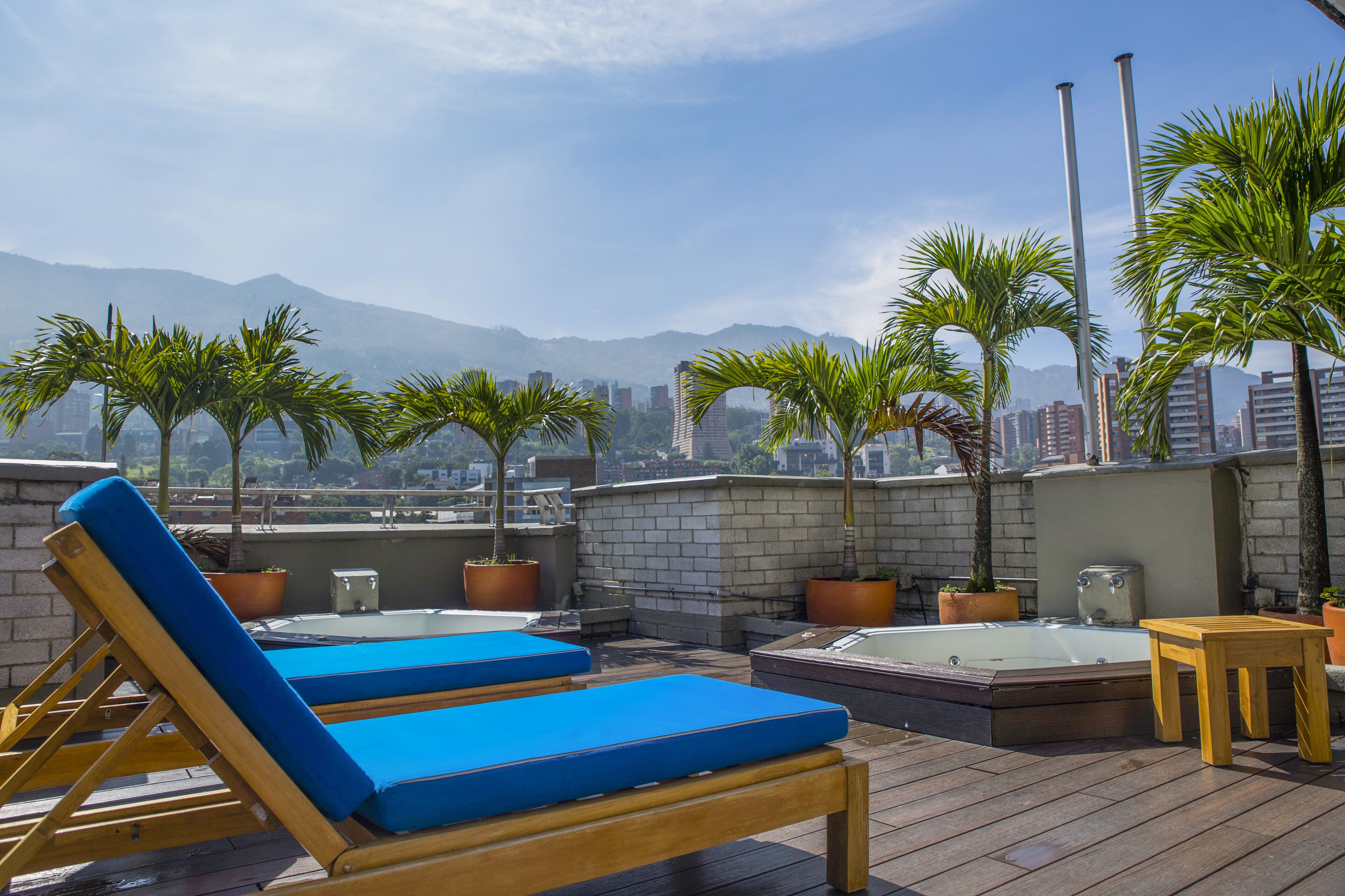 A rooftop lounge with lounge chairs and a hot tub, surrounded by potted plants and overlooking a cityscape with mountains in the background.