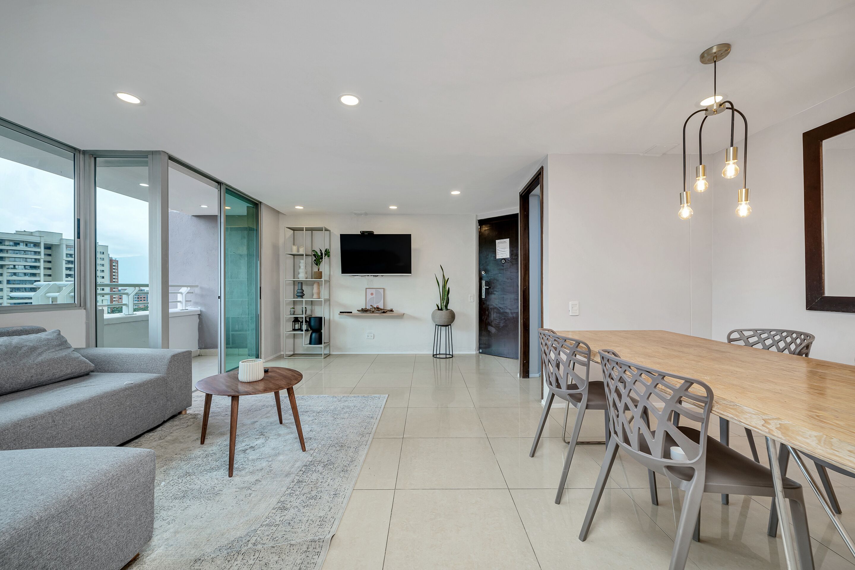 A modern living room with a sofa, coffee table, and dining table with chairs. There is a TV mounted on the wall and a plant on a side table.