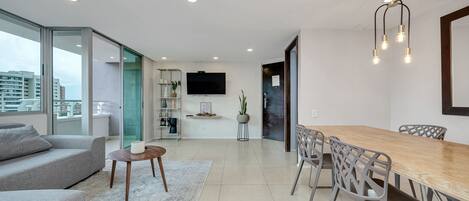 A modern living room with a sofa, coffee table, and dining table with chairs. There is a TV mounted on the wall and a plant on a side table.