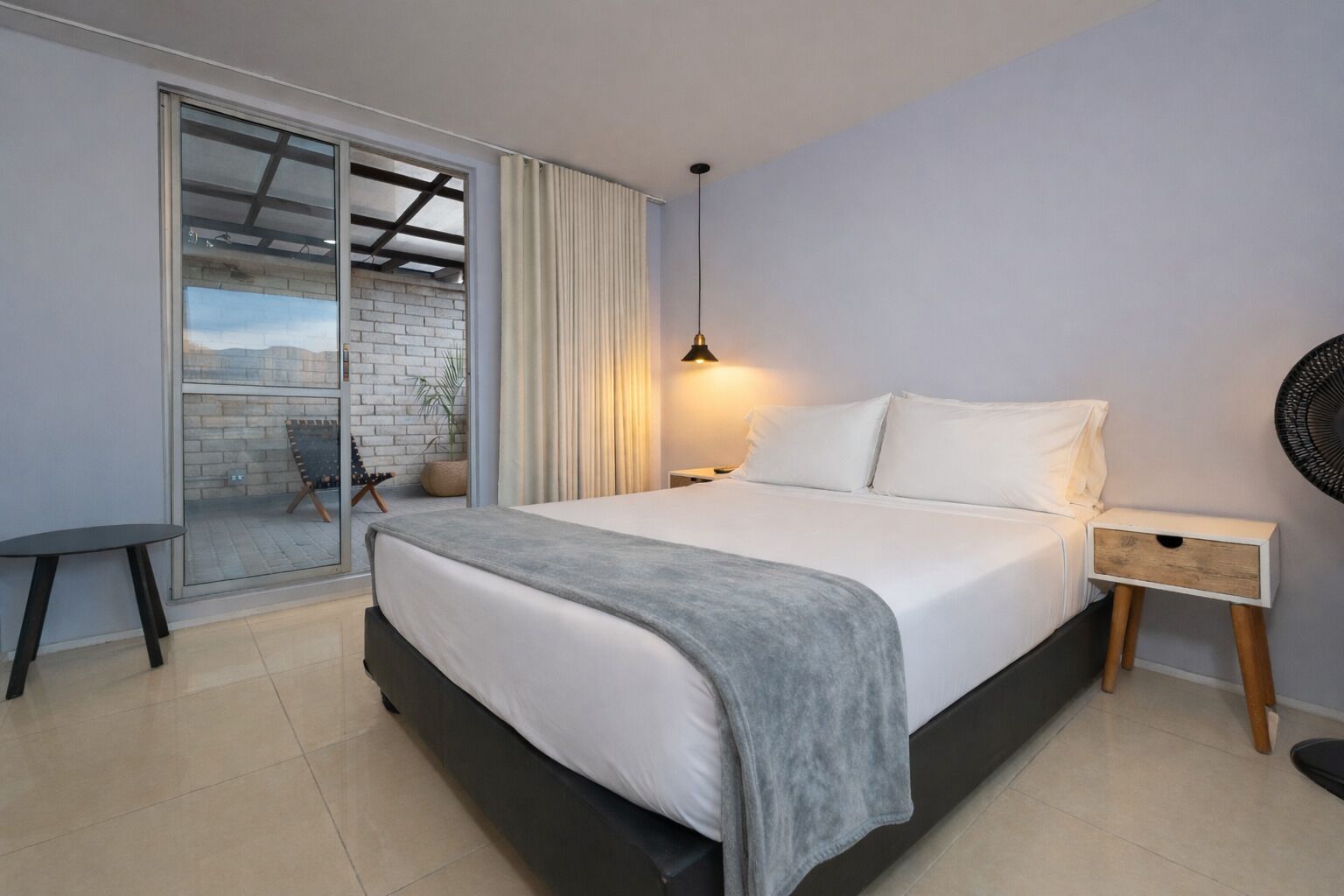 Quarto com cama, mesa de cabeceira, abajur, cadeira e vista por uma porta de vidro.