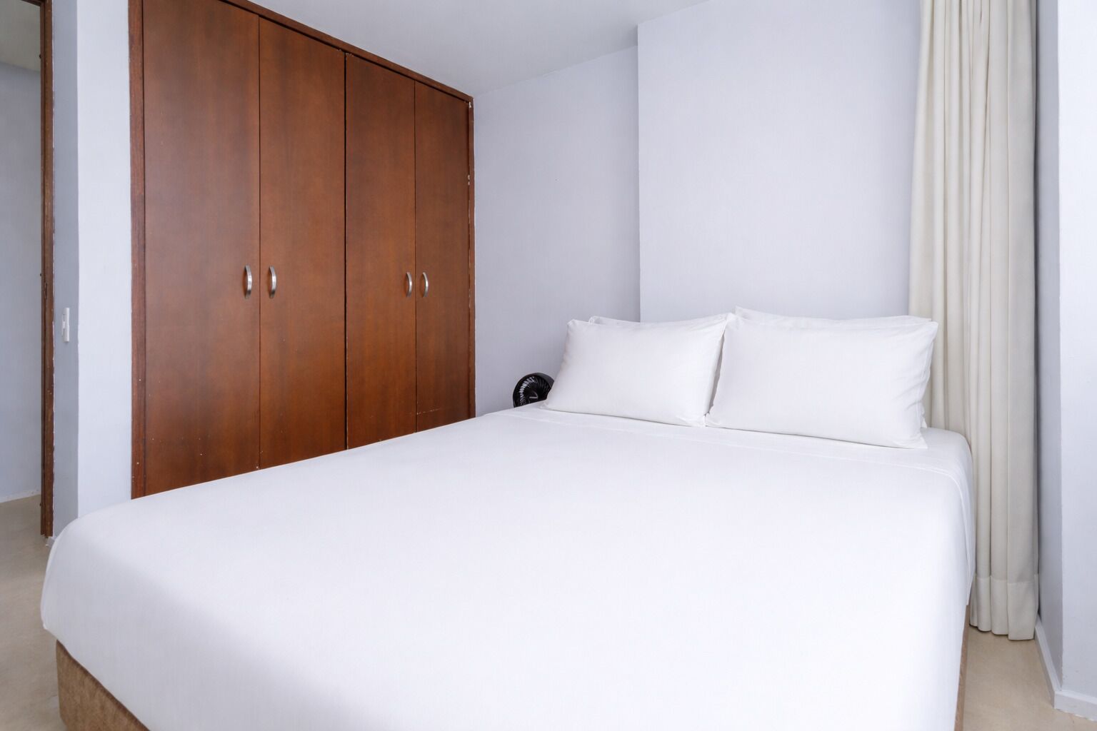 A bedroom with a large bed, white bedding, a wooden wardrobe, and a fan.