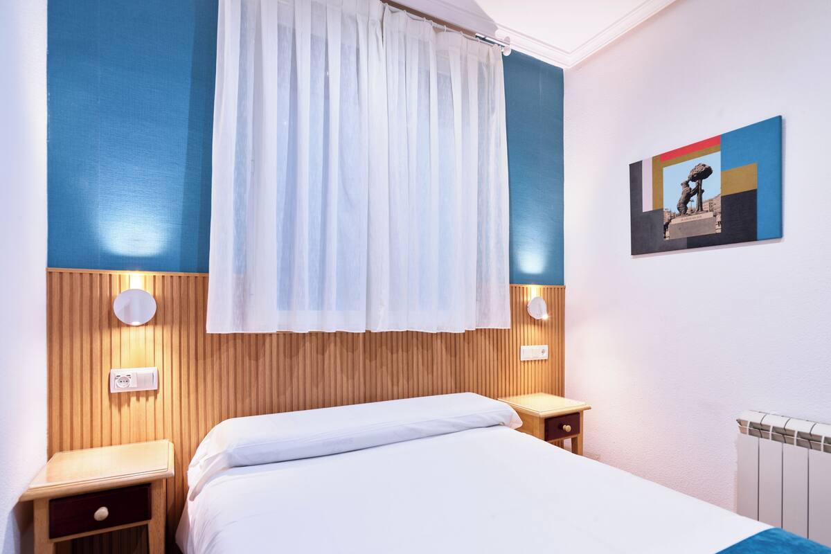 A hotel room with a bed, wooden headboard, white curtains, bedside tables, and a wall-mounted light fixture.