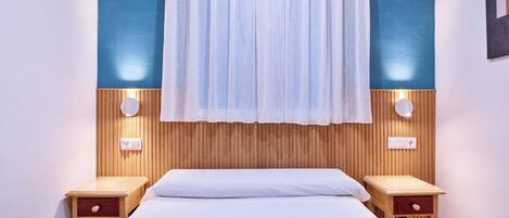Une chambre d’hôtel avec un lit, deux tables de chevet, une lampe fixée au mur et une fenêtre avec un rideau léger.