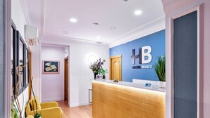 Een hotel receptie met een houten receptiebalie, een blauwe muur met het logo van het hotel, een ingelijste bloemillustratie en een geel zitgedeelte.