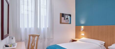 A hotel room with two beds, a desk with a chair, a window with sheer curtains, and a wall-mounted picture.