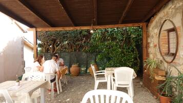 An outdoor patio with wooden tables and white plastic chairs, surrounded by potted plants and a brick wall.