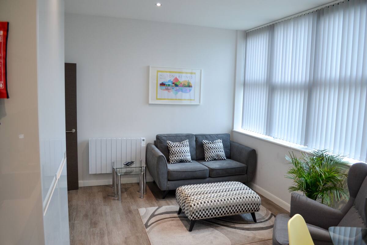 A modern living room with a grey sofa, patterned ottoman, and a framed artwork on the wall.