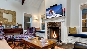 A cozy living room with a fireplace, plaid chairs, a coffee table with playing cards, and a large TV screen displaying a skier.