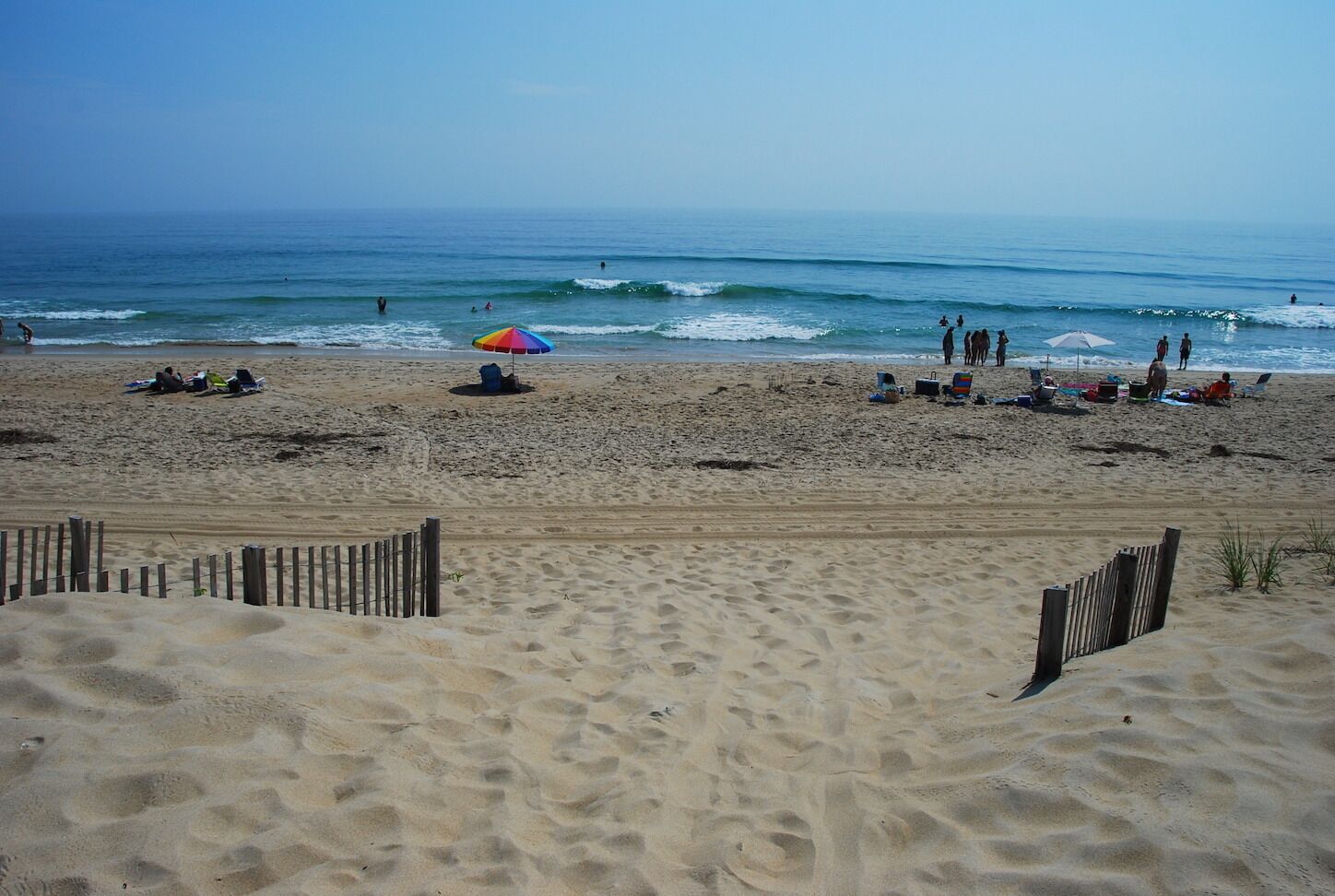 Une plage de sable avec une clôture en bois au premier plan, quelques personnes et des chaises de plage au milieu, et l’océan en arrière-plan.