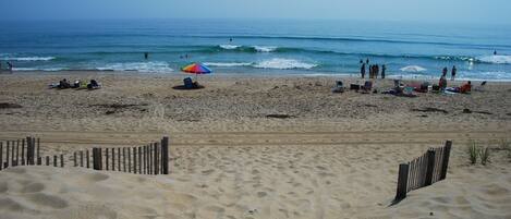 Une plage de sable avec une clôture en bois au premier plan, quelques personnes et des chaises de plage au milieu, et l’océan en arrière-plan.
