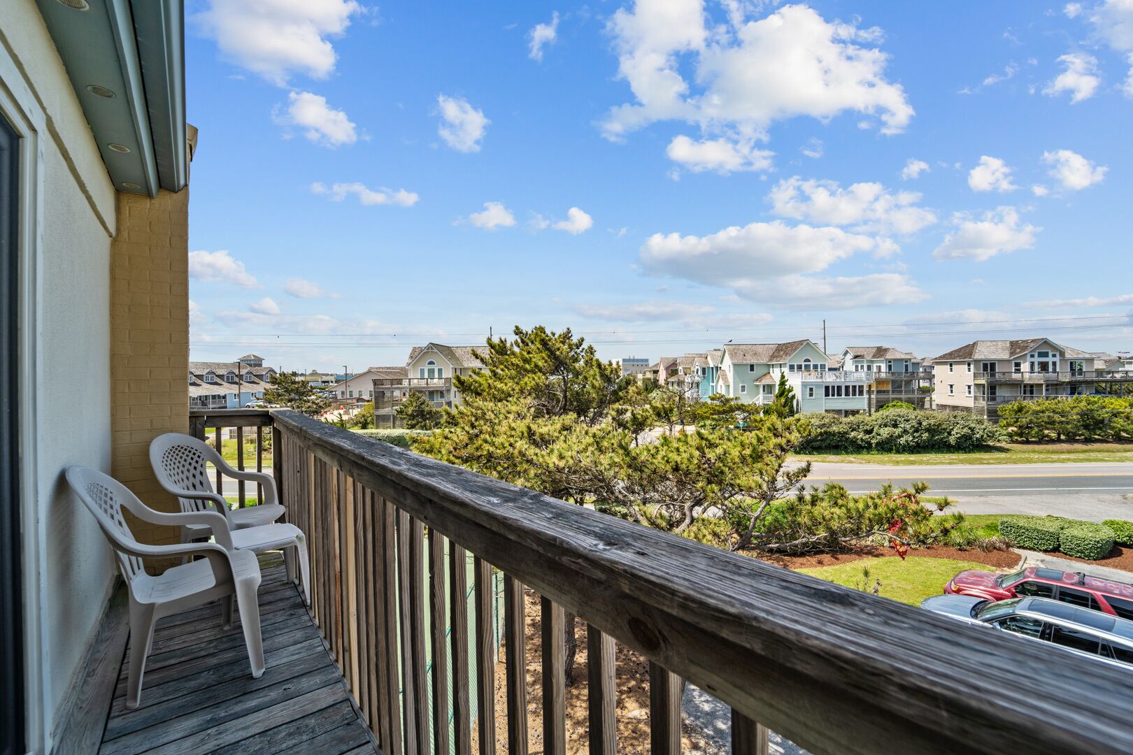 A wooden balcony with two white plastic chairs, overlooking a row of townhouses and a road with parked cars.