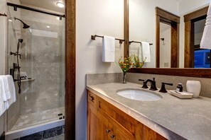 A bathroom with a glass-enclosed shower, a sink with a marble countertop, wooden cabinets, and a large mirror.