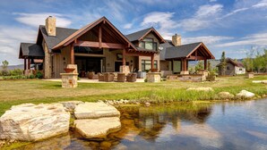 A large, modern house with a stone exterior, a large patio, and a pond in the foreground. - Abode at Twilight Ranch (Oakley)
