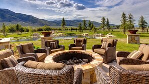 A circular outdoor seating area with wicker furniture and a central fire pit, surrounded by a scenic landscape with mountains and a pond. - Abode at Twilight Ranch (Oakley)